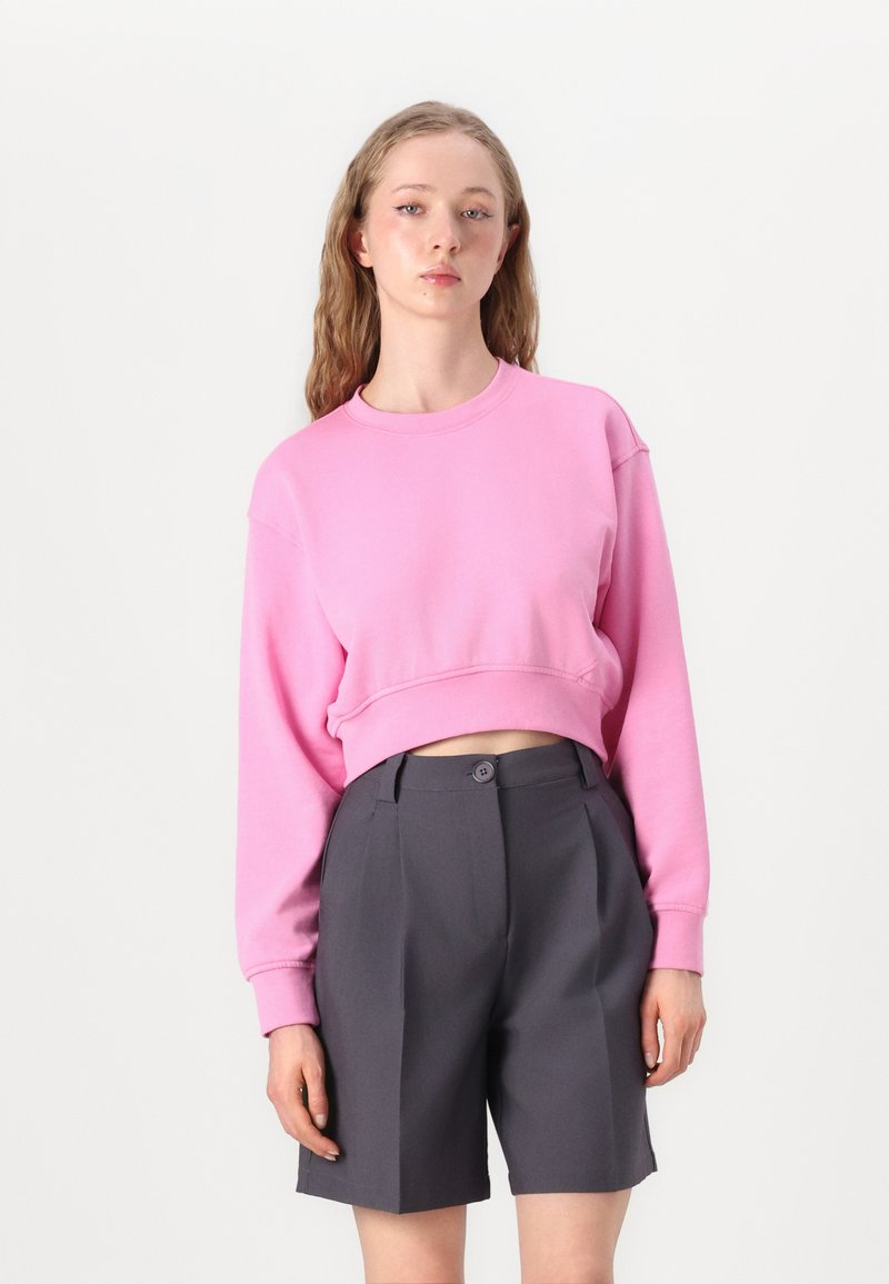 Fuksia kort sweatshirt med lange ermer, rund hals og ribbestrikket kant, kombinert med skreddersydde sortgrå shorts med knappelukking.