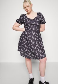 Vestido preto com mangas curtas em forma de balão, apresentando uma estampa de borboleta branca, decote quadrado e barra com evasê. Sapatos pretos e meias brancas.