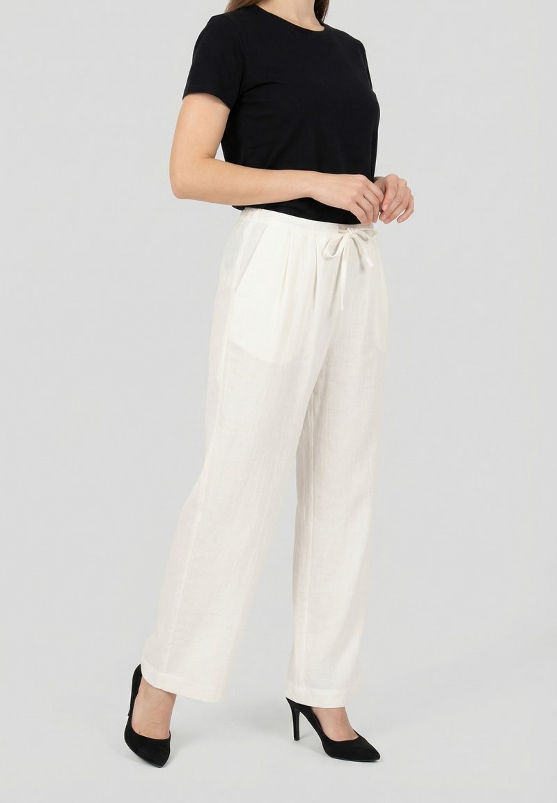 Femme portant un haut noir à manches courtes, un pantalon blanc taille haute avec cordon de serrage, et des talons noirs à bout pointu, debout devant un fond gris.