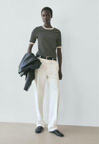 Frau trägt ein schwarz-weiß gestreiftes Hemd, weiße Hose und schwarze Schuhe, hält in der linken Hand eine schwarze Lederjacke.