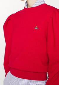 Suéter rojo con mangas abullonadas, puños y dobladillo acanalados. Presenta un diseño bordado de una corona y un planeta en tonos dorado, azul y verde.