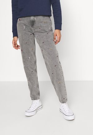 Osoba nosząca szare jeansy acid-wash z małymi haftowanymi logo oraz białe wysokie trampki, stojąca na jednolitym tle.