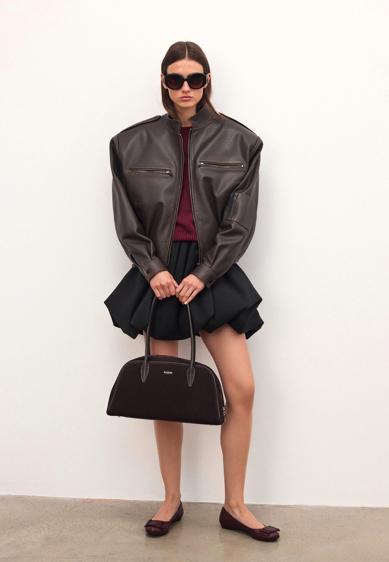 Smeđa kožna oversized jakna s patentnim zatvaračima, bordo pulover, crna suknja s voluminoznim krojem, smeđa torba i tamne balerinke.