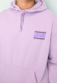 Lila huvtröja gjord av mjukt material, med en ficka fram och en grafisk logotyp med geometriska detaljer på bröstet.