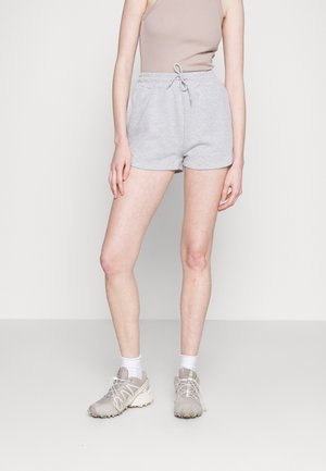 Personne portant un débardeur beige, un short gris clair à cordon, des chaussettes blanches et des chaussures de sport beiges, debout sur un fond neutre.