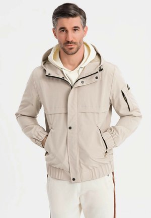 Mężczyzna w beżowej kurtce z kapturem na białym hoodie, stojący z rękami w kieszeniach na tle białego tła.