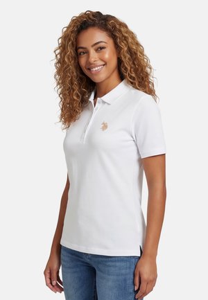 Smiling woman with curly hair wearing a white short-sleeve polo shirt with a small embroidered logo and blue jeans, standing sideways.
