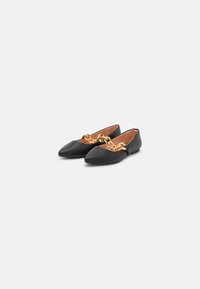 Zapatos planos negros con punta afilada, con un acento de cadena dorada en la parte frontal. Hechos de cuero con una textura suave.