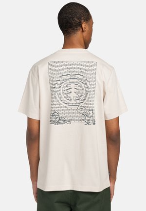 Camiseta de algodón color crema con un diseño gráfico que representa una pared de ladrillos, con personajes de dibujos animados y burbujas de diálogo en la parte posterior.