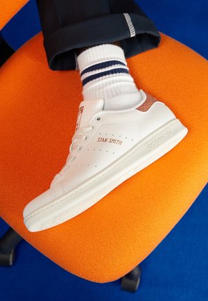 Białe trampki Adidas Stan Smith noszone z białymi skarpetkami z niebieskimi paskami, spoczywające na pomarańczowym krześle na tle niebieskiej podłogi.