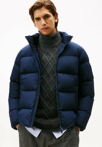 Doudoune bleu marine avec des sections matelassées, col haut et fermeture éclair, portée sur un pull col roulé texturé gris et un pantalon à motifs.