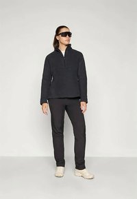 Črn flis pulover z zadrgo na ovratniku, kombiniran s črnimi hlačami in svetlimi čevlji. Model nosi temna sončna očala. Preprost, sodoben dizajn.