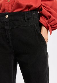 Pantalon en velours côtelé noir avec une surface texturée, comportant une fermeture à bouton à la taille et des poches latérales. Accentuée par un haut ample.