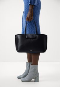 Borsa a tracolla in pelle nera con un accento in tessuto intrecciato sulla parte anteriore. Presenta manici lunghi e una forma rettangolare. Texture liscia.