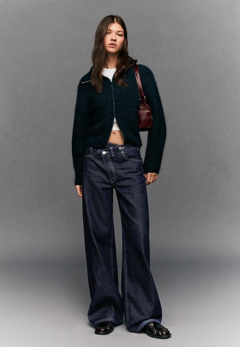 Tamnozeleni pleteni kardigan na patentni zatvarač sprijeda, bijeli kratki top, široke tamnoplave traperice s ravnim nogavicama i mala bordo torba. Uže crne ravne cipele.