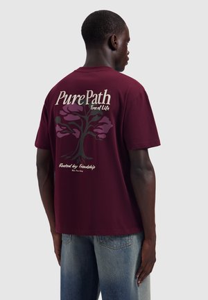 Man met zijn gezicht van de camera af, draagt een bordeauxrood T-shirt met een boomgrafiek en tekst "PurePath Tree of Life Rooted by Friendship" op de achterkant.