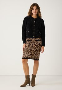 Eine Frau trägt eine schwarze, geknöpfte Strickjacke, einen Rock mit Leopardenmuster und Taschendetails sowie braune Wildleder-Stiefeletten und steht vor einem schlichten Hintergrund.