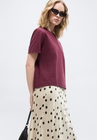 Femme portant des lunettes de soleil noires style œil de chat, un haut manches courtes bordeaux, une jupe crème à pois noirs, tenant un sac à main noir.