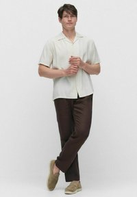 Chemise à manches courtes de couleur beige clair avec un devant boutonné, associée à un pantalon marron foncé. Les chaussures sont des mocassins beige à finition texturée.