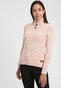 Maglione a maglia rosa con colletto alto, tre bottoni marroni, polsini e orlo a costine. Presenta un motivo texture e un'etichetta del marchio di lato.