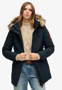 Navy blue parka with a faux fur hood, zip front, and side pockets. Thick knit cream sweater underneath, paired with light blue jeans.