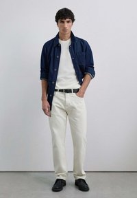 Chaqueta de mezclilla, camiseta blanca, pantalones color crema, cinturón negro y zapatos negros. El modelo está de pie con una mano en el bolsillo, fondo neutro.