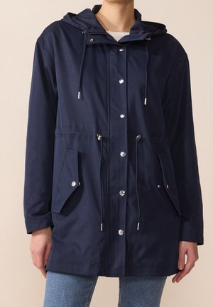 Marineblauwe regenjas met capuchon, gemaakt van een gladde, waterdichte stof. Beschikt over zilveren hardware, trekkoorden en voorzakken.