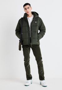 Tumši zaļa pūkainā jaka ar izšūtu dizainu, rāvējslēdzēju un kapuci. Valkāta virs baltas krekla, apvienota ar olīvzaļām kordūras biksēm un baltām kedām.