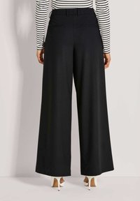 Vrouw in zwarte wijde broek, gestreepte lange mouw top en witte hoge hakken, staand tegen een neutrale achtergrond.