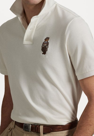 Polo shirt de culoare deschisă cu un țesut texturat, având un detaliu brodat cu un urs și o guler clasic. Purtați cu pantaloni bej și un brâu maro.
