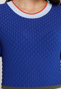 Pull bleu tricoté avec un motif texturé, doté d'un col côtelé bleu clair et rouge et d'un ourlet inférieur vert.