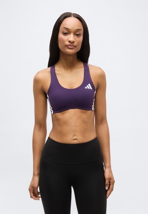 Žena s dlhými tmavými vlasmi, oblečená v fialovom športovom podprsenke s logom a čiernych legínach s vysokým pásom, stojaca pred jednoduchým pozadím.
