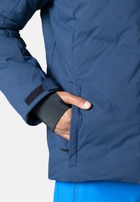 Marineblaue isolierte Jacke mit weicher Textur, ausgestattet mit einem schwarzen Bündchenfutter und einem Klettverschlussriemen am Ärmel. Enthält ein seitliches Taschen-Detail.