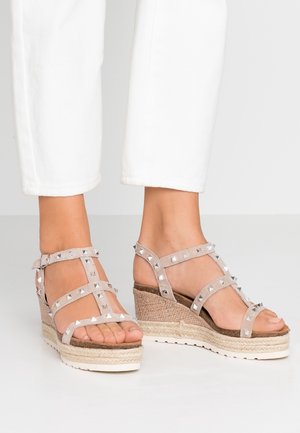 Fødder iført beige kilehælssandaler med sølvnitter og ankelremme, kombineret med hvide knælange bukser på en hvid gulvflade.