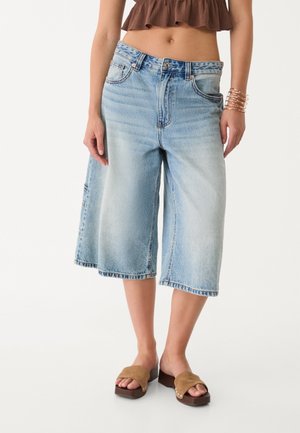 Femme portant un short large en jean bleu clair, un top court marron et des sandales marron ouvertes sur fond uni.