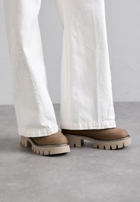 Chaussures à plateforme marron avec une tige texturée, une semelle claire contrastante et des encoches prononcées. Portées avec un pantalon blanc à jambes larges.