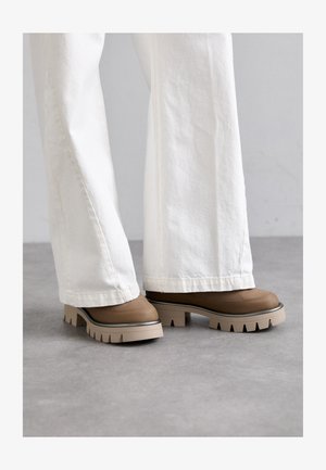 Brune plateau-sko med en tekstureret overdel, en kontrastfuld lys sål og markante takker. Båret med hvide bukser med vidde ben.