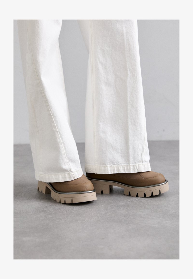 Chaussures à plateforme marron avec une tige texturée, une semelle claire contrastante et des encoches prononcées. Portées avec un pantalon blanc à jambes larges.