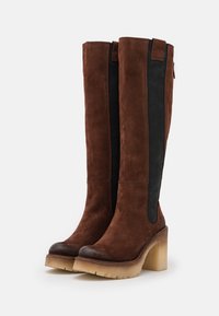 Botas de ante marrón hasta la rodilla con paneles elásticos negros en los lados y un tacón grueso de bloque beige. Presentan una punta redonda lisa y una suela texturizada.