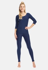 Donna in piedi a piedi nudi che indossa un top blu navy con maniche a tre quarti e leggings abbinati, sorridente con una mano sul fianco.