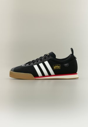 Zwarte Adidas Samba sneaker met witte strepen, rubberen zool, rood streepdetail en gouden logo aan de zijkant, getoond in zijaanzicht op een neutrale achtergrond.