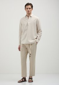 Helle beige Langarmbluse mit Kragen und drei Knöpfen, kombiniert mit hellbeigen Jogginghosen mit Kordelzug und braunen Sandalen. Einfaches Design.