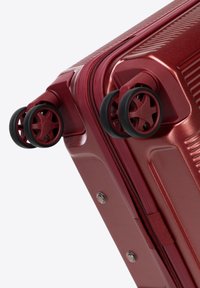Valigia rigida rossa con superficie testurizzata, dotata di quattro ruote nere e chiusura a zip. Finitura liscia e design resistente.