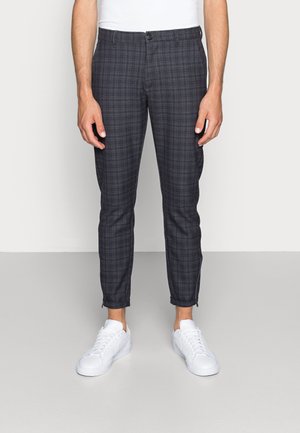 Homme portant un pantalon slim gris foncé à carreaux et des baskets blanches, debout devant un fond clair uni.