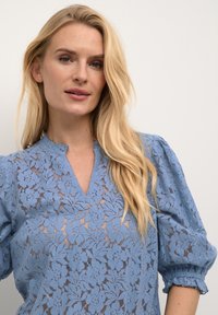 Blusa azzurro chiaro in pizzo con motivo floreale, scollatura a V e maniche a sbuffo con polsini elastici. Dettagli del design testurizzati visibili.