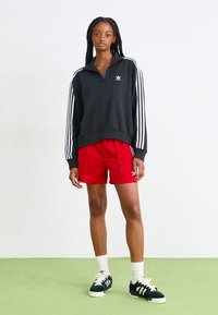 Juoda zip-up džemperis su baltais dryžiais, derinamas su raudonomis šortais. Modelis vilki juodus ir baltus sportinius batelius, demonstruodamas sportišką, kasdienį stilių.