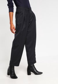 Svarta plisserade byxor med en lös passform, tillverkade av ett texturerat tyg, i kombination med spetsiga svarta ankelstövlar.