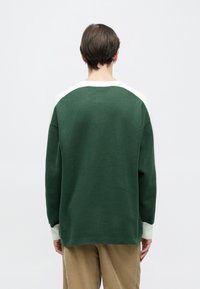 Zöld kötött pulóver fehér gallérral és ujjal. Laza szabású és texturált felületű. Hátulról varrási részletek láthatók.