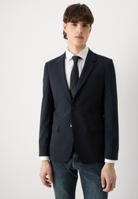 Kerge kangaga mereväe sinine bleiser, millel on üks nööp, sakilised kraed ja kaks esikotit, kantud valge särk ja must lips.