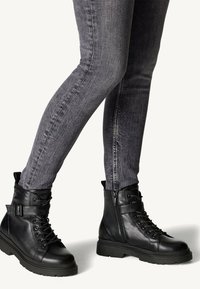 Bottes de combat en cuir noir avec lacets et fermeture éclair sur le côté, semelle en caoutchouc et détail de sangle à la cheville, associées à un jean gris ajusté.